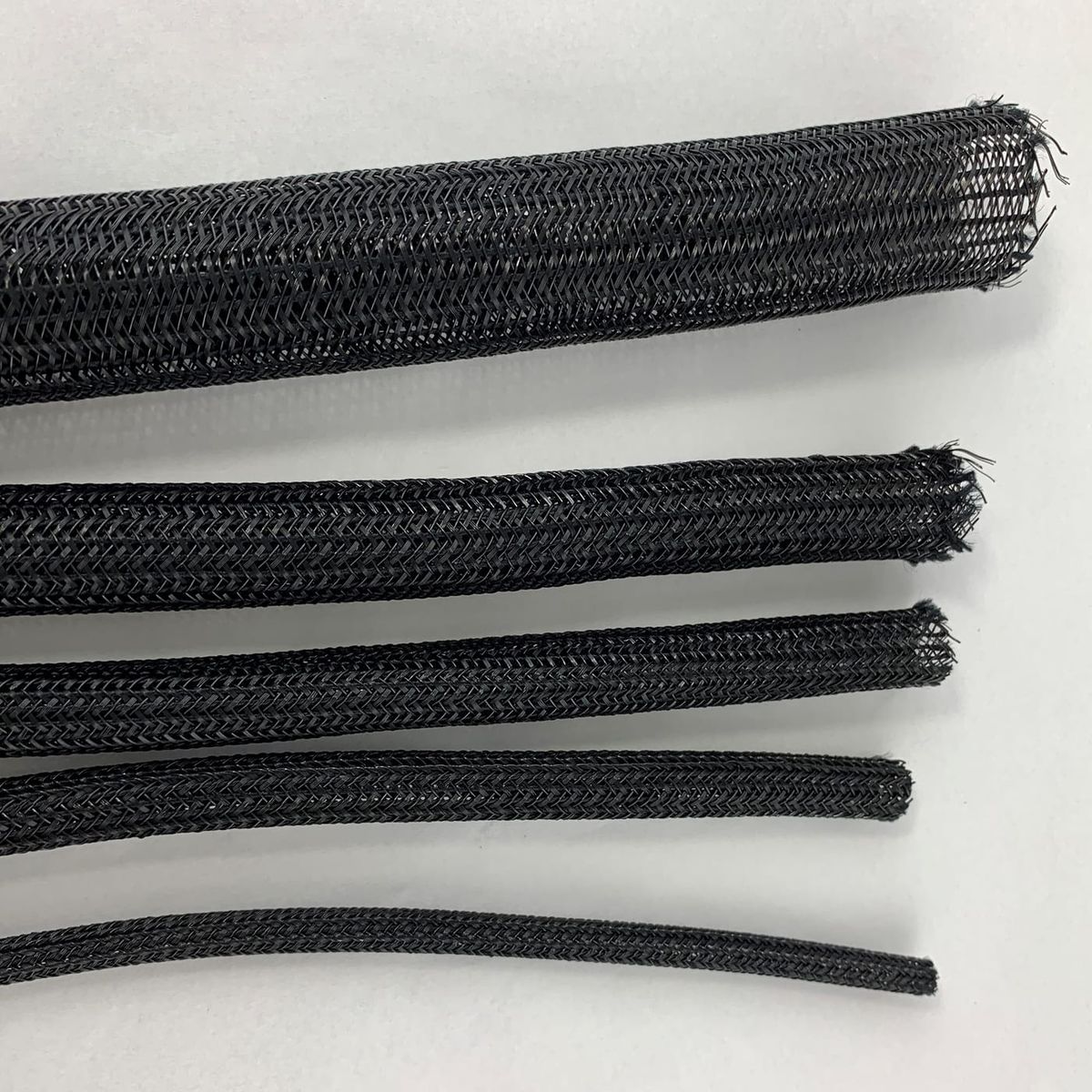 Five diameters of black braided sleeve laid side by side on a white surface, each cut end left open to show the braid structure