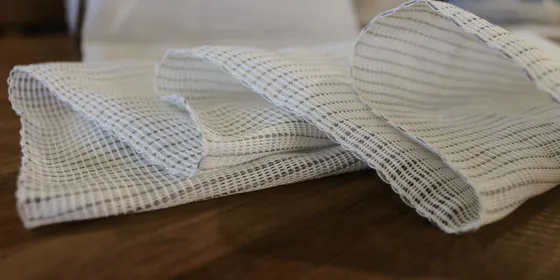 Folded lengths of white open-mesh heating fabric on a dark wooden bench, the grey conductive yarn visible running through the weave