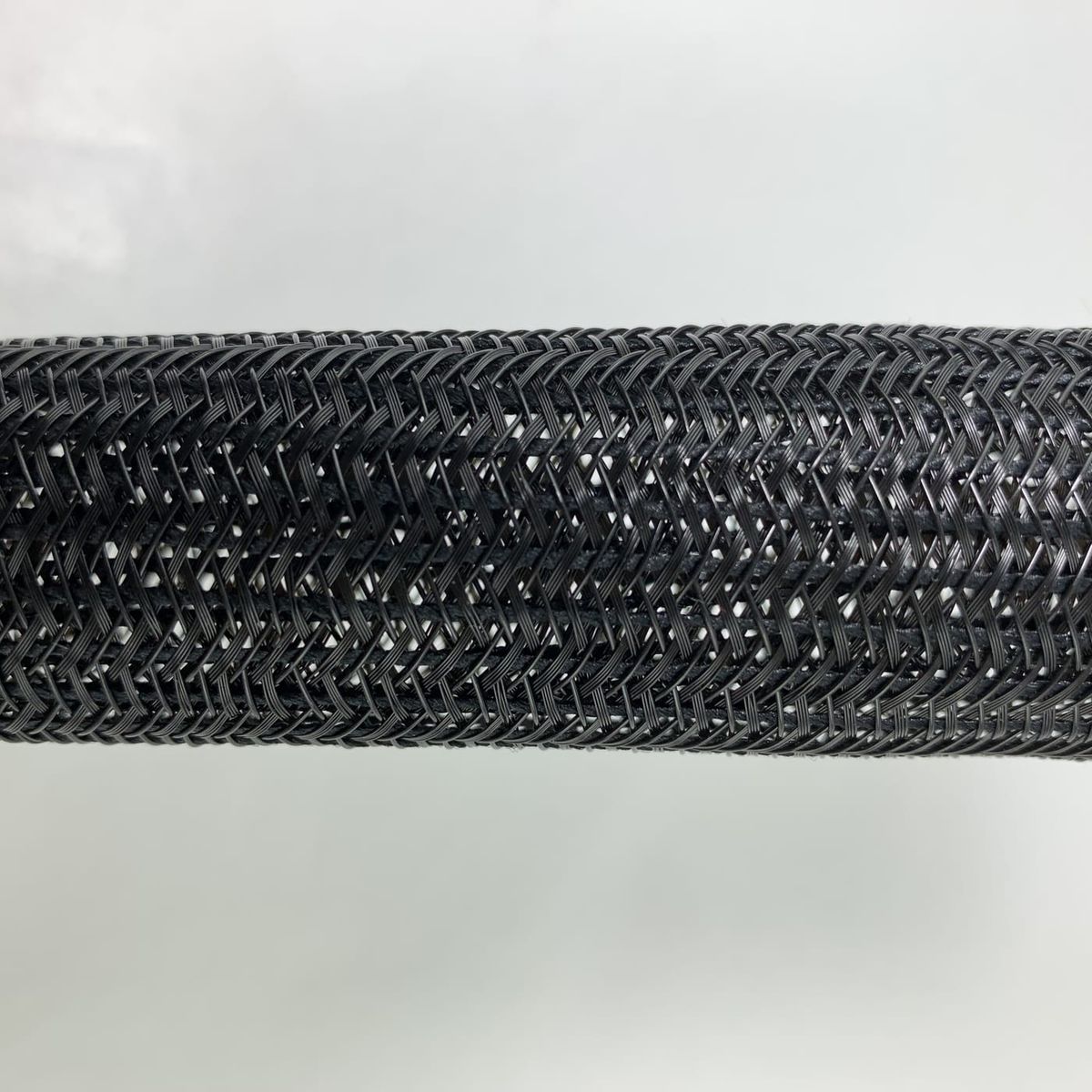 Macro photograph of a black braided tube running horizontally across the frame, its surface woven in a diagonal herringbone pattern of crossing filament bundles with small diamond-shaped gaps between them, against an out-of-focus pale background